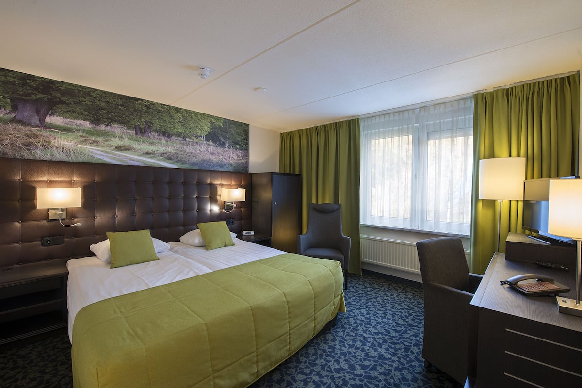 A hotel room with a large bed, a desk with a telephone, a chair, and a painting on the wall.