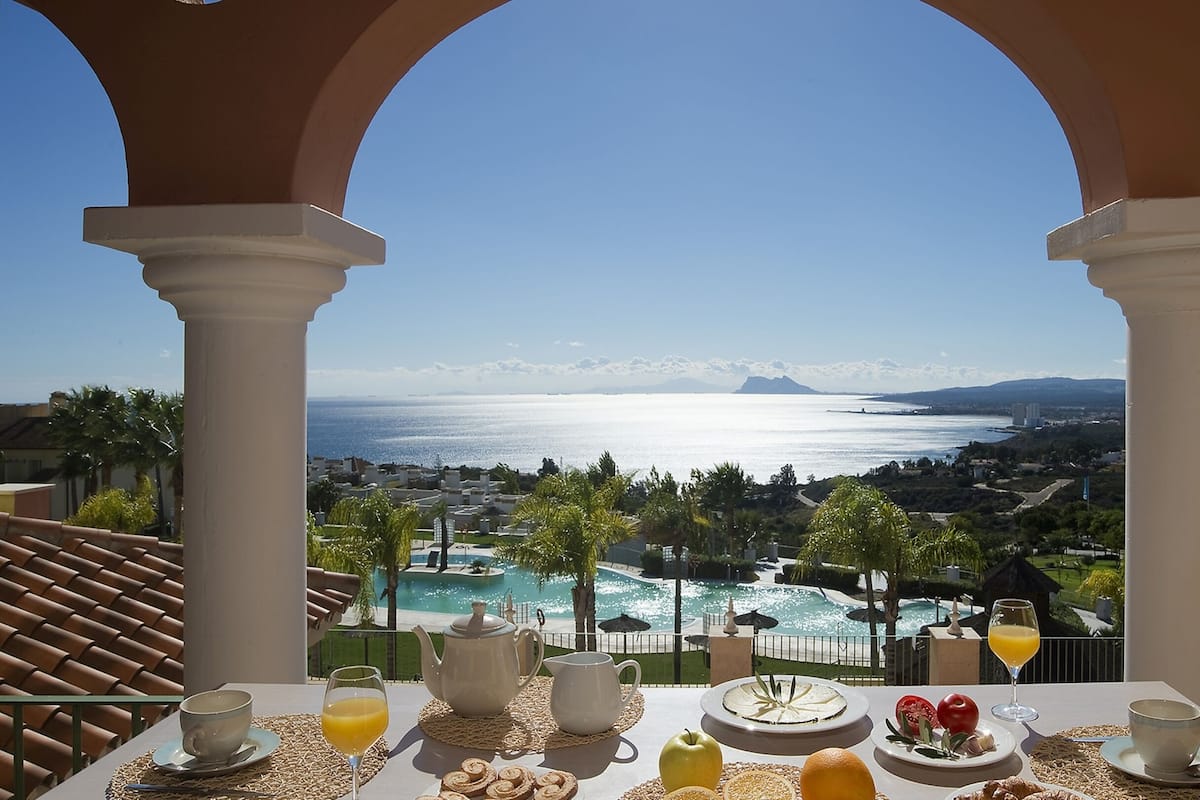 A table with breakfast items and a view of a coastal resort with a pool and mountains in the distance.