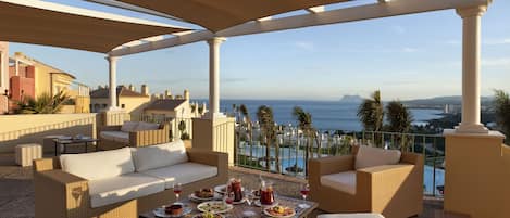 Ein überdachter Außenbereich mit Korbmöbeln, einem Tisch mit Tellern und Gläsern sowie Blick auf das Meer und Gebäude.