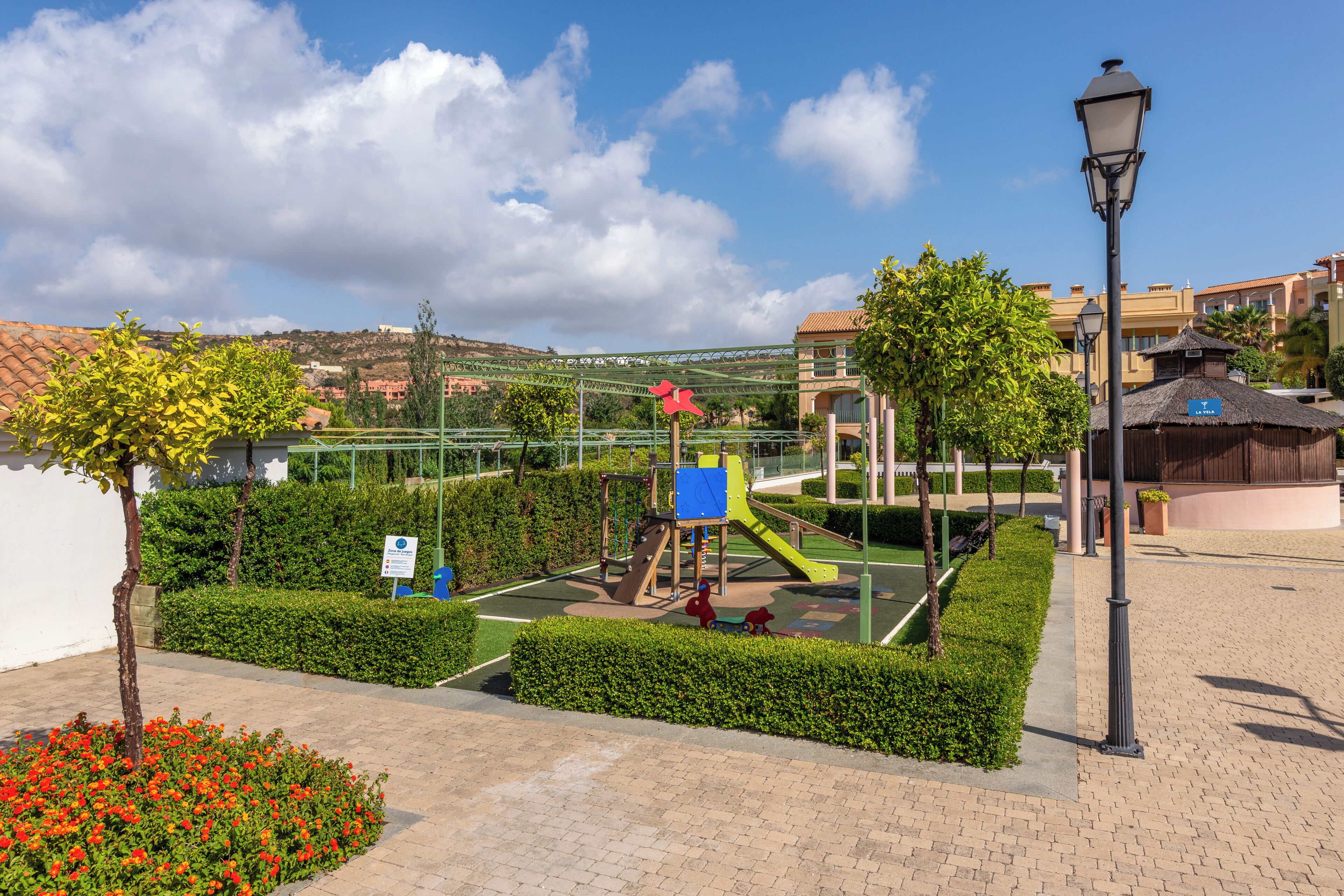 Ein Spielplatz mit Rutschen und Klettergerüsten, umgeben von ordentlich geschnittenen Hecken und einem gepflasterten Weg.