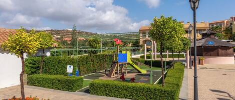 Ein Spielplatz mit Rutschen und Klettergerüsten, umgeben von ordentlich geschnittenen Hecken und einem gepflasterten Weg.