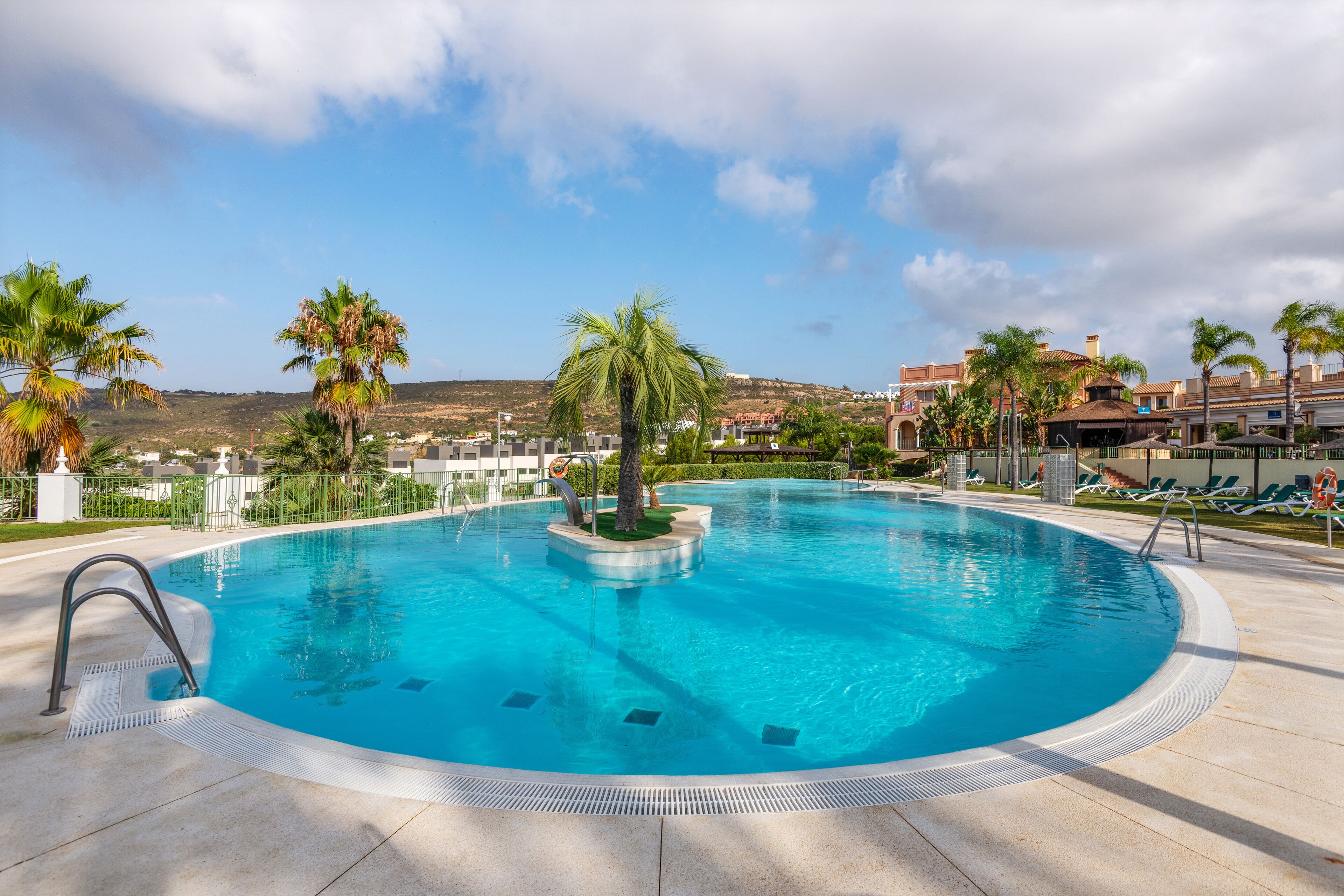 A circular swimming pool with a palm tree in the center, surrounded by lounge chairs and a view of buildings in the background.