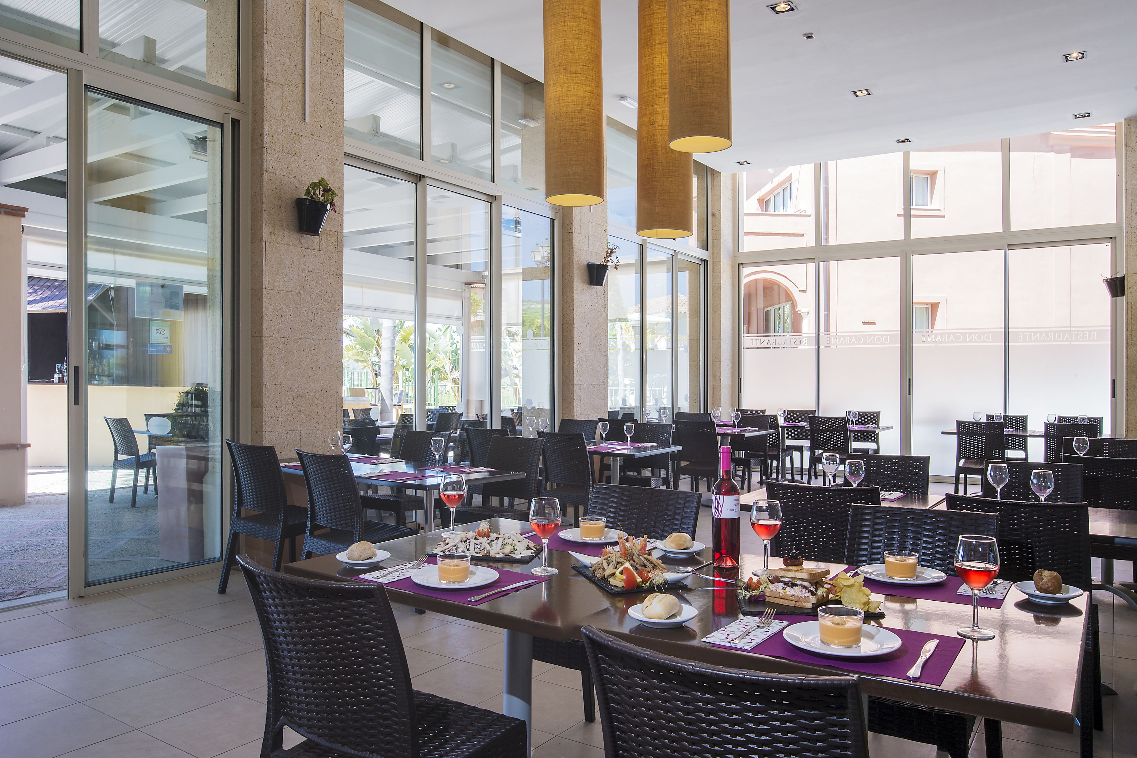 A spacious restaurant with multiple tables and chairs, large windows, and hanging pendant lights.