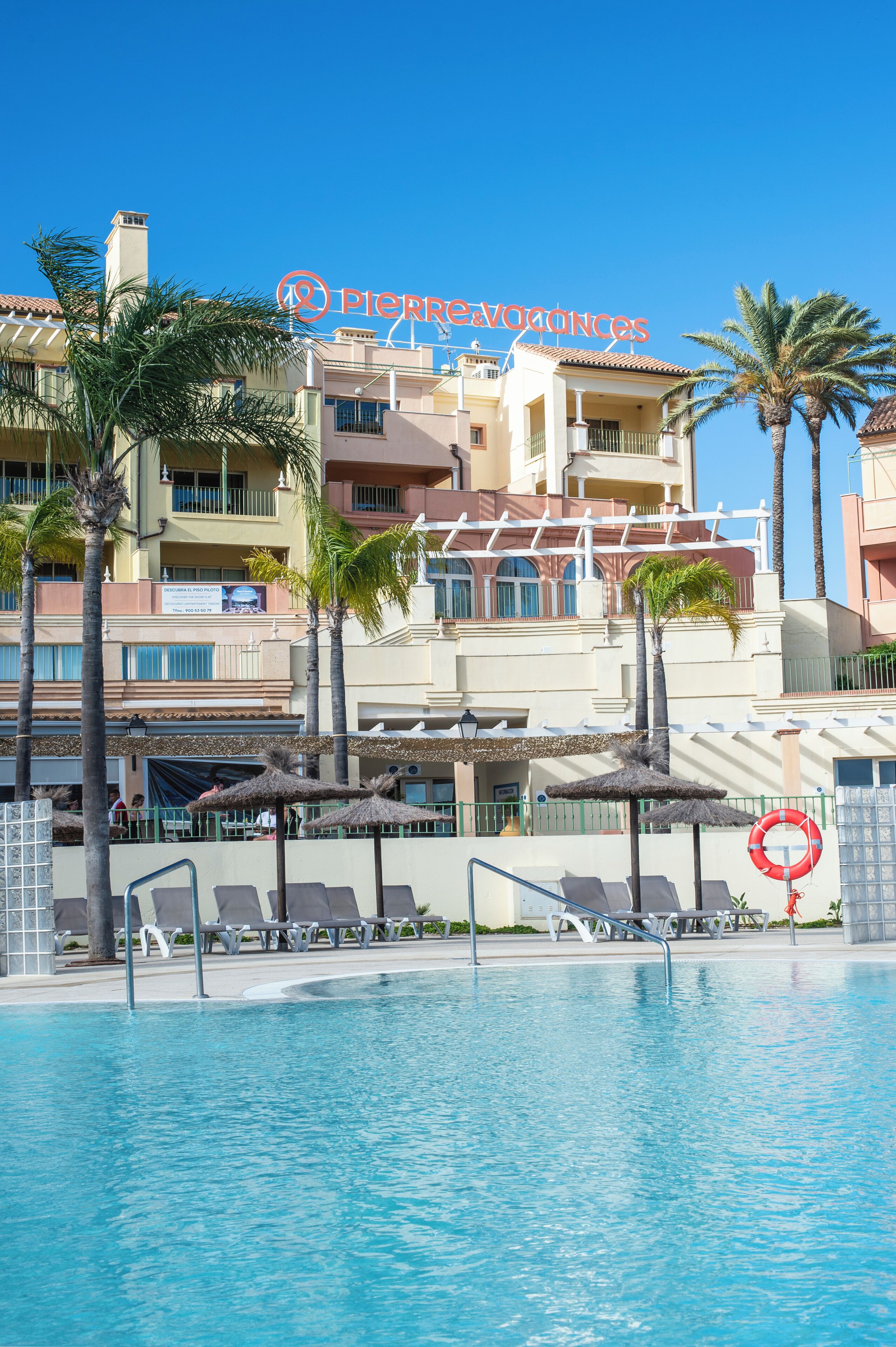 A hotel pool area with lounge chairs, palm trees, and a lifebuoy.