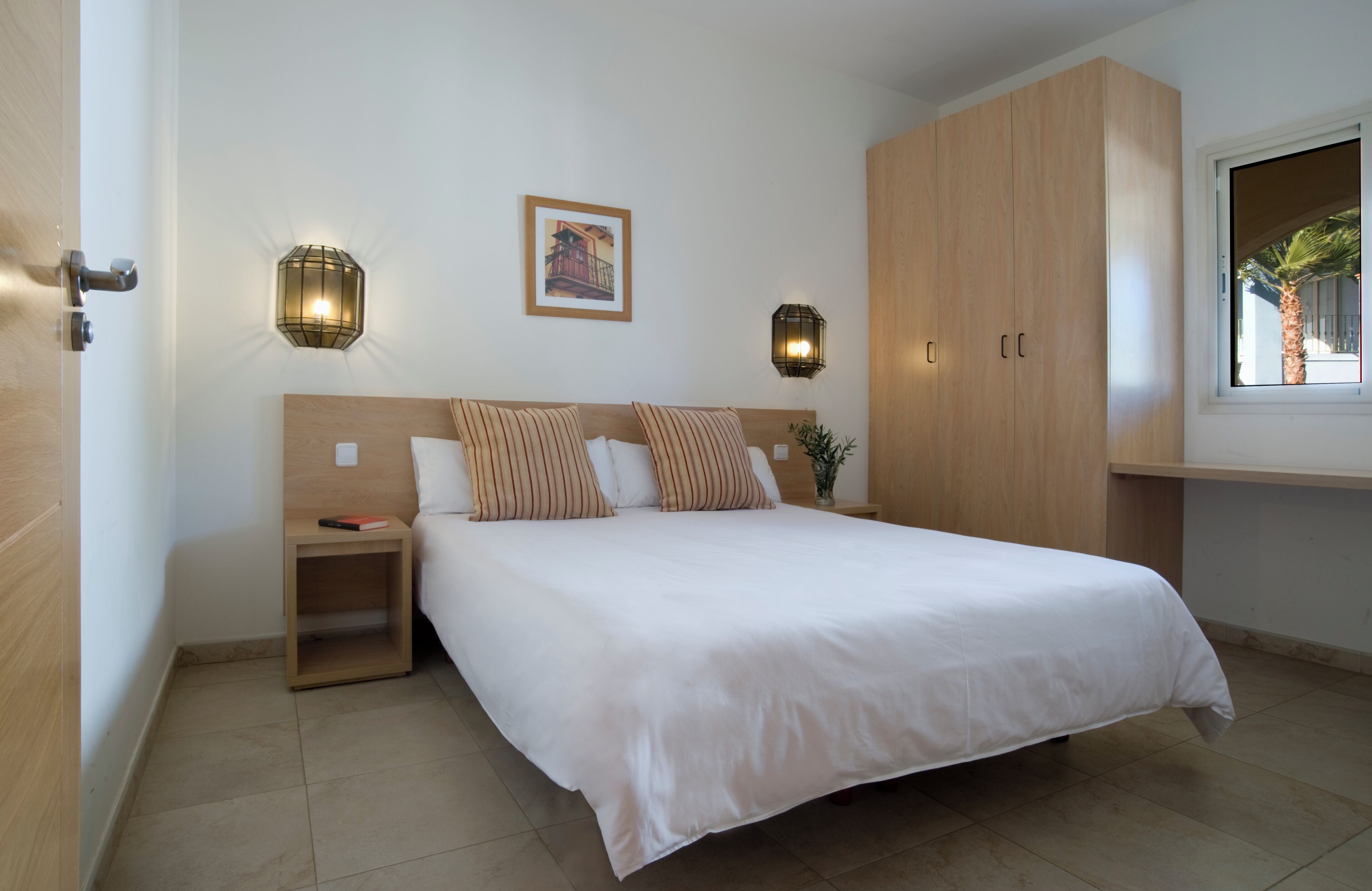 Ein ordentlich eingerichtetes Schlafzimmer mit einem großen Bett, einem Holzschrank, Nachttischlampen und einem Fenster mit Blick auf Palmen.