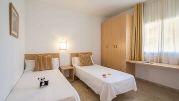 A hotel room with two single beds, a wooden wardrobe, a nightstand with a lamp, and a window with curtains.