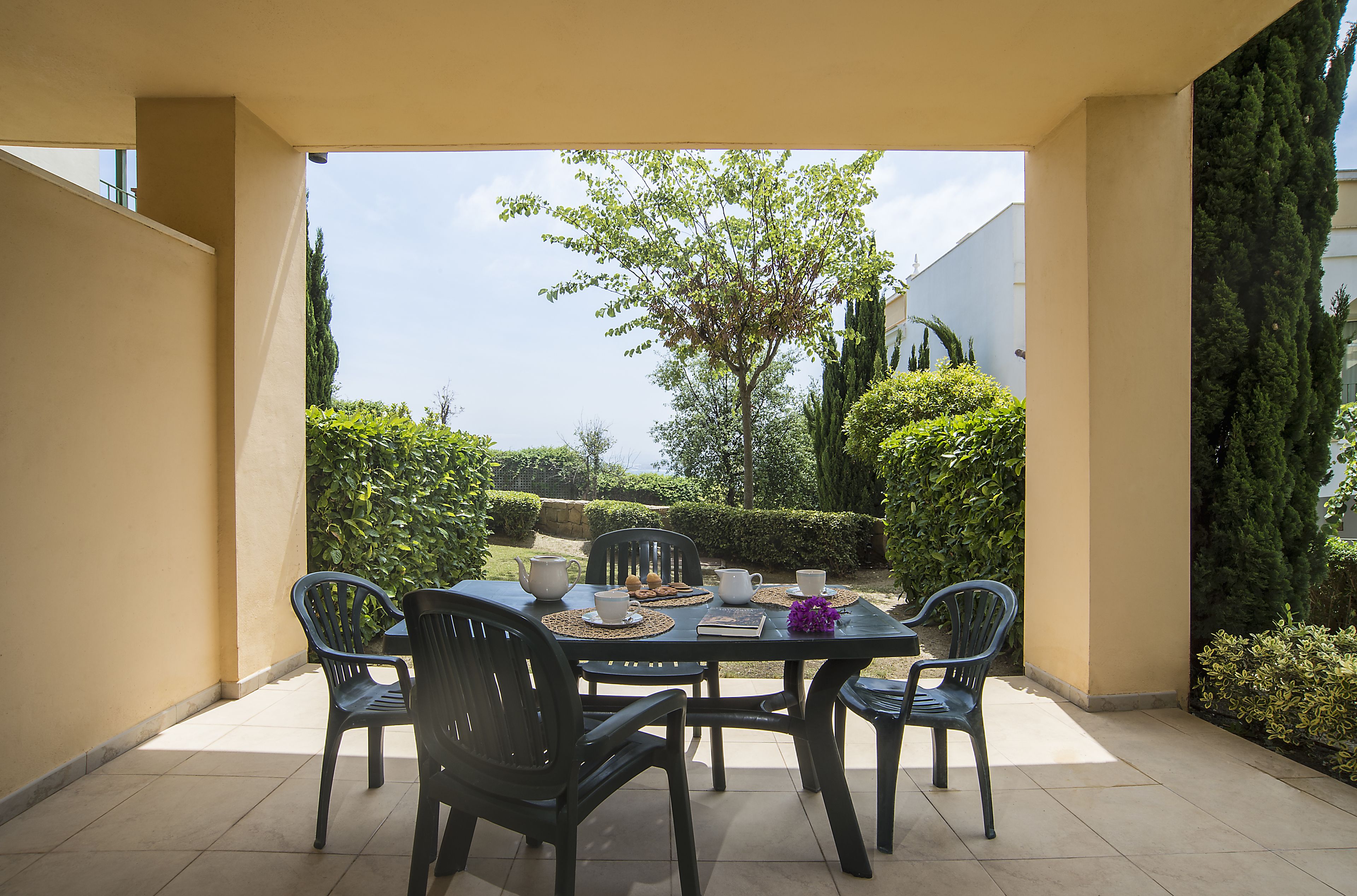 A covered patio with a table set for four, surrounded by chairs, and a view of a garden with hedges and trees.