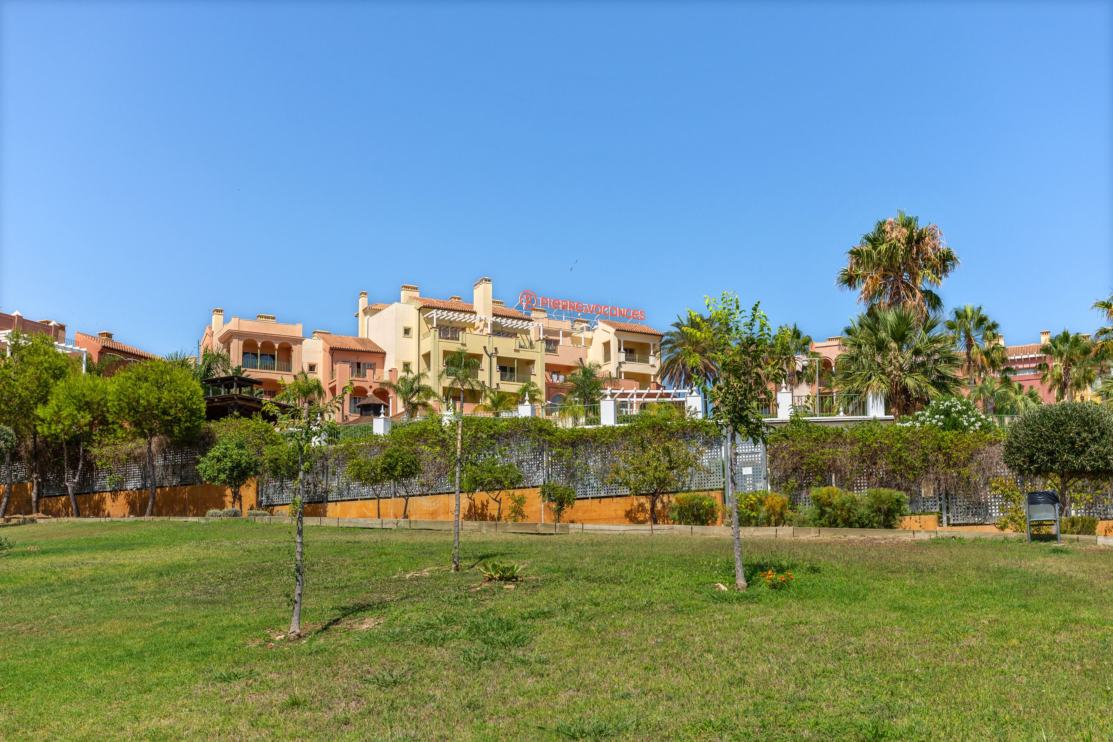 A hotel with a large, open grassy area in the foreground, a fence, and a variety of trees.
