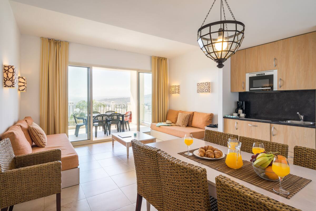 A modern hotel room with a dining area, kitchen, and a balcony with a view.