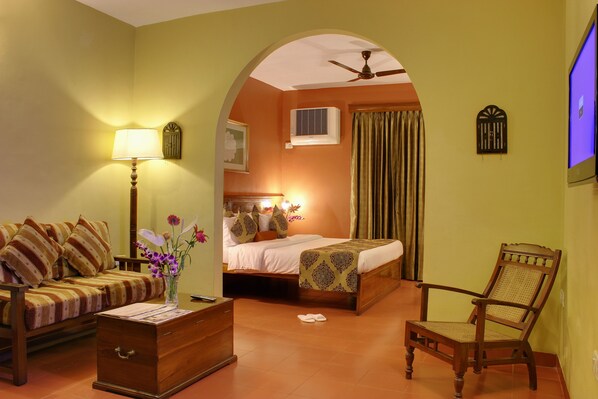 A hotel room with a sofa, a wooden chest, a chair, a bed, and a television. - Pride Sun Village Resort & Spa (Arpora)