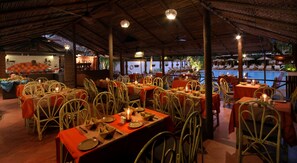 An indoor dining area with tables set for guests, a bar in the background, and a pool visible through the windows. - Pride Sun Village Resort & Spa (Arpora)