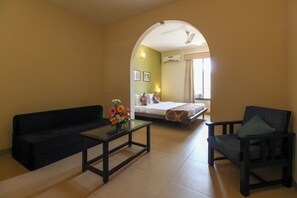 A hotel room with a sofa, armchair, and a small table with flowers. There is a bed with a patterned blanket and pillows. - Pride Sun Village Resort & Spa (Arpora)