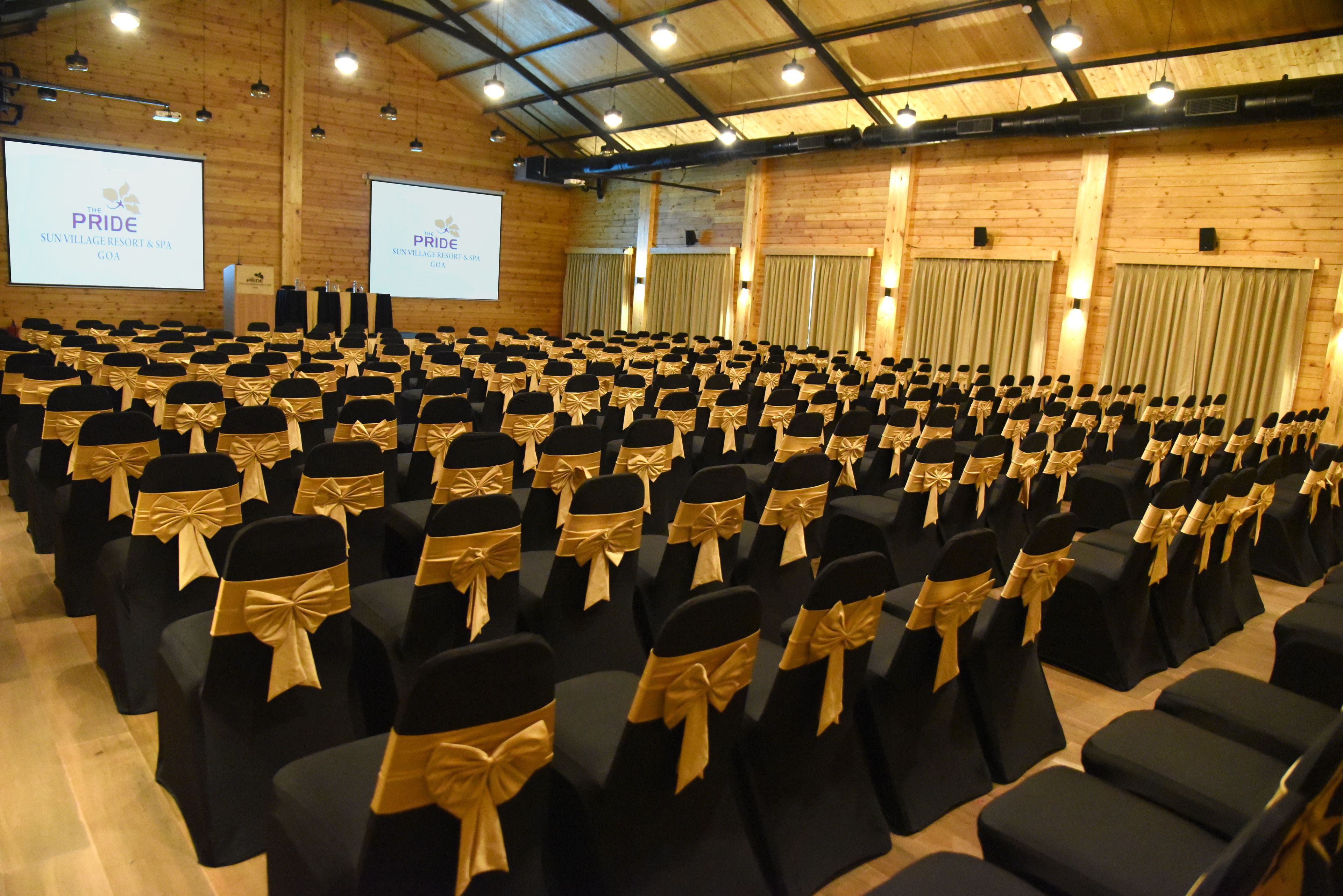 A conference room with rows of black chairs with gold sashes, wooden walls, and a projector screen displaying "PRIDE SUNVILLAGE RESORT & SPA GOA".