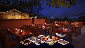 An outdoor dining area with multiple tables set for a meal, surrounded by wooden chairs and tables, under a large tree. - Pride Sun Village Resort & Spa (Arpora)