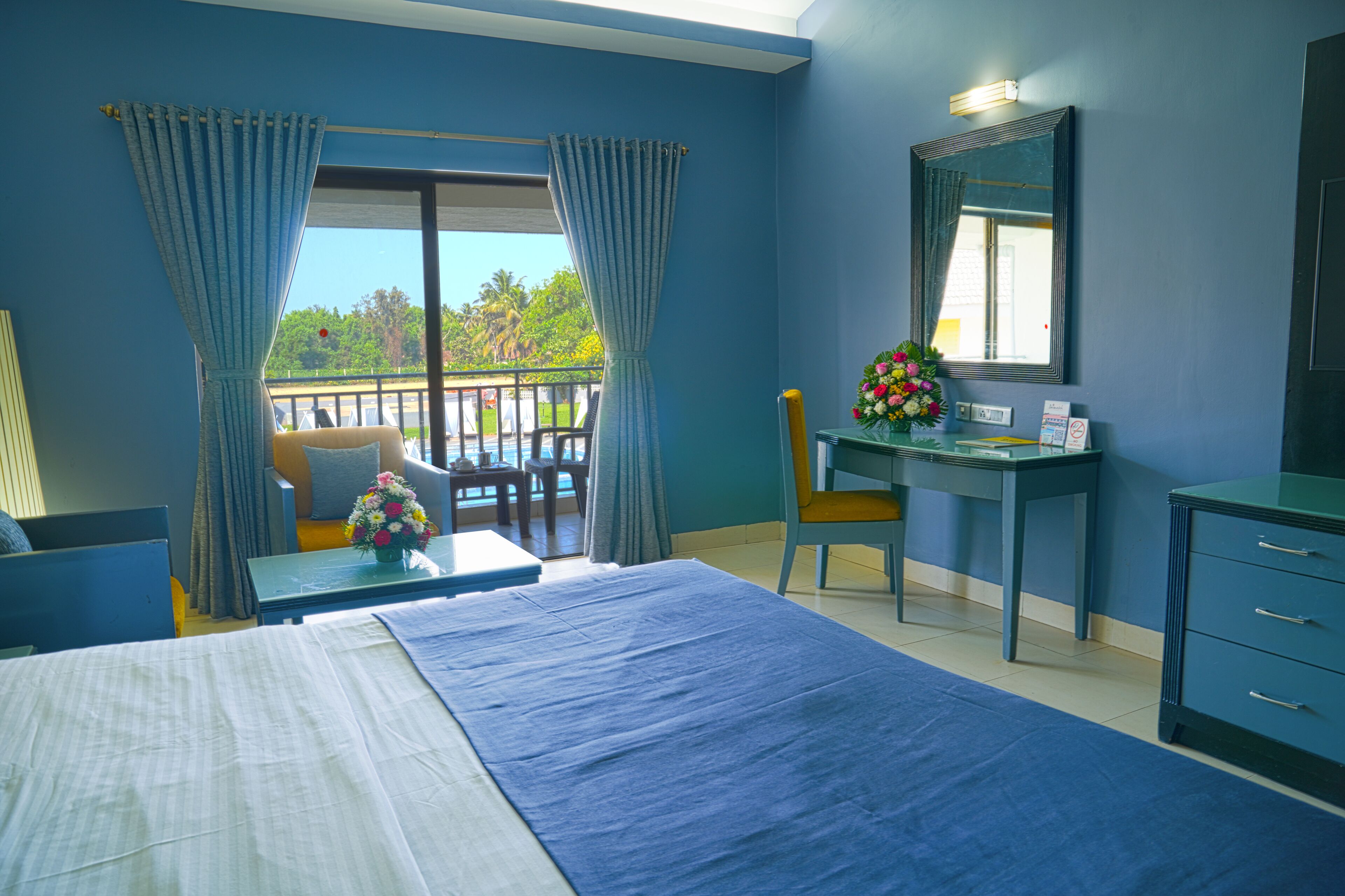 A hotel room with a bed, desk, and chair, a balcony with a view, and a mirror on the wall.