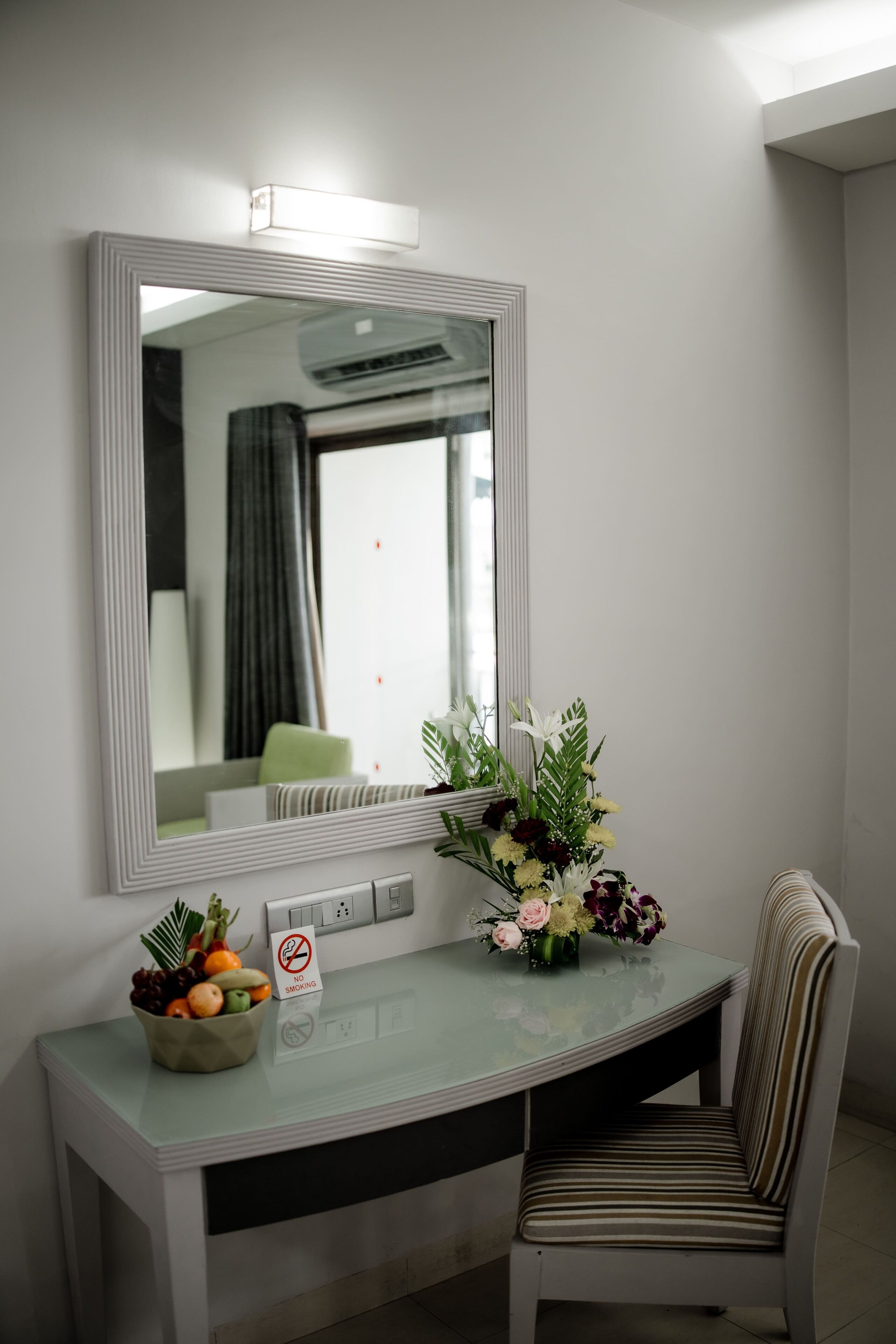 A hotel room with a large mirror, a small desk with a no-smoking sign, a chair, and a vase of flowers.