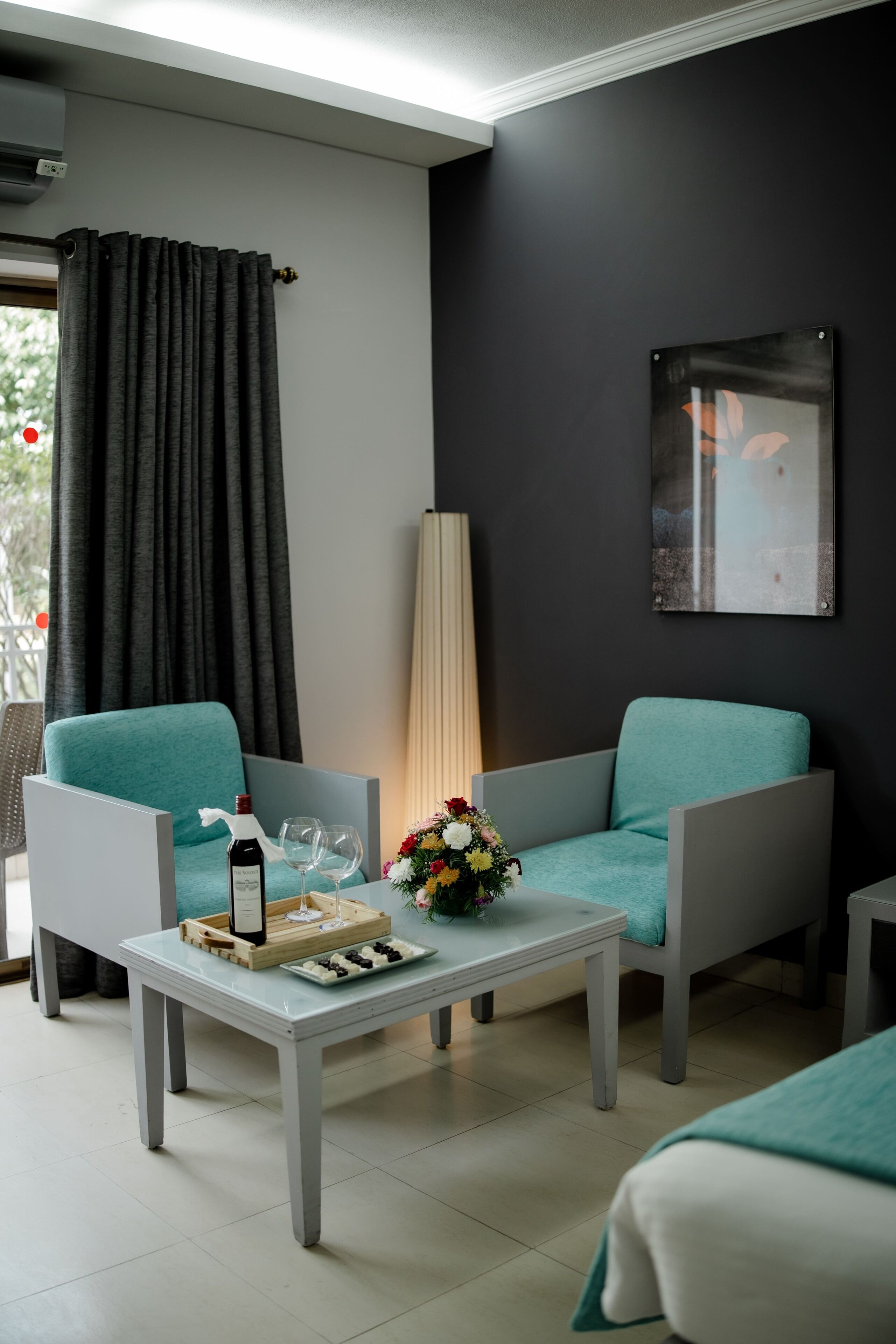 Una habitación de hotel moderna con dos sillones color turquesa, una mesita pequeña con una botella y vasos, y un jarrón de flores.