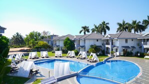 A swimming pool with lounge chairs and a small waterfall feature, surrounded by palm trees and a building with a tiled roof. - Regenta Baywatch Resort Goa (Colva)