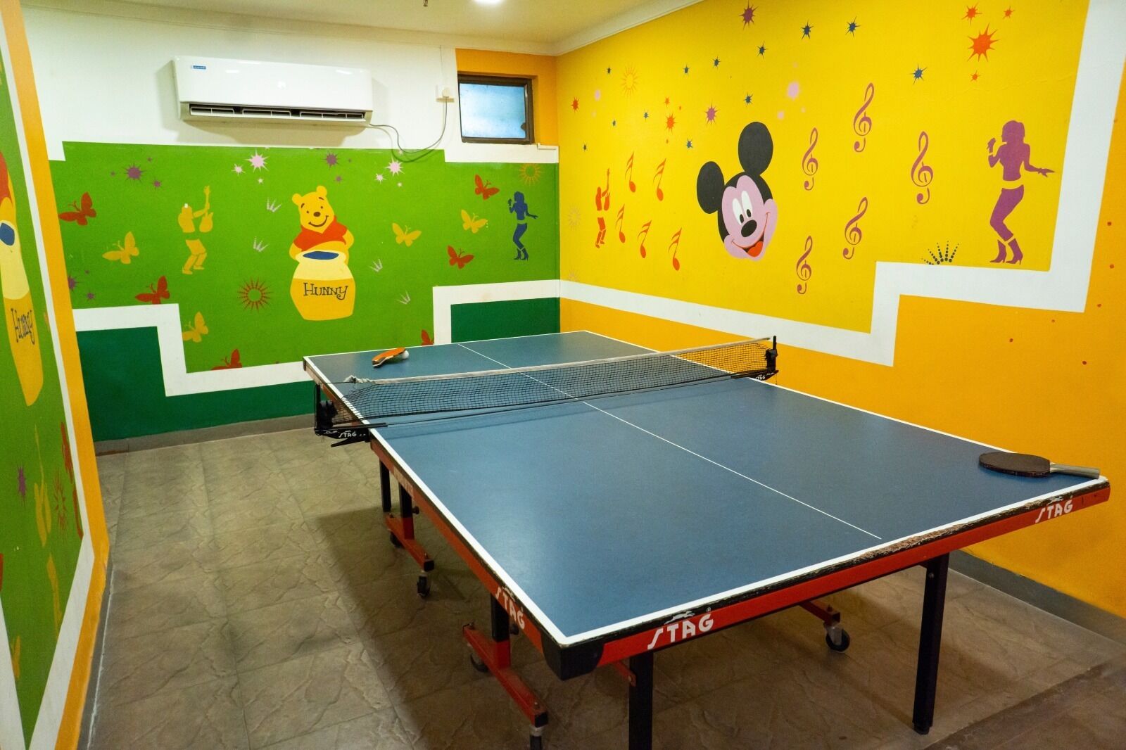 Una mesa de ping pong en una habitación con paredes de colores, una ventana y un equipo de aire acondicionado.