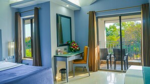 Una habitación de hotel con una cama, un escritorio con una silla, una mesita con un florero y una ventana con cortinas, además de un balcón con sillas.
