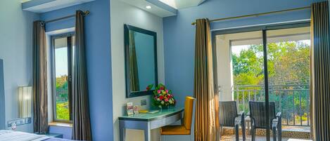 A hotel room with a bed, a desk with a chair, a small table with a vase of flowers, a window with curtains, and a balcony with chairs.