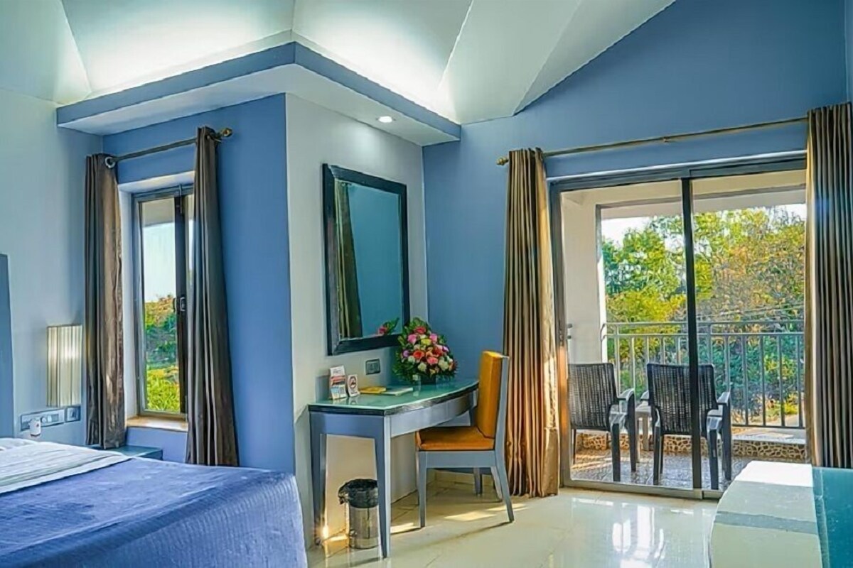 A hotel room with a bed, a desk with a chair, a mirror, a window with curtains, and a balcony with chairs.