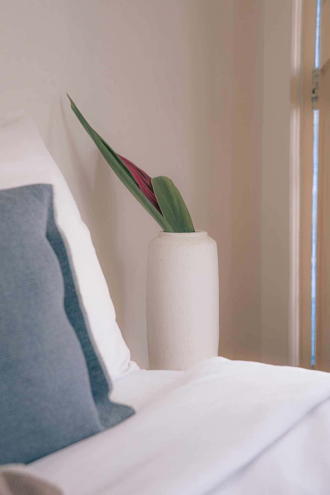 Ein Bett mit weißer Bettwäsche und einem blauen Kopfkissen, eine weiße Vase mit Pflanze sowie eine Tür im Hintergrund.