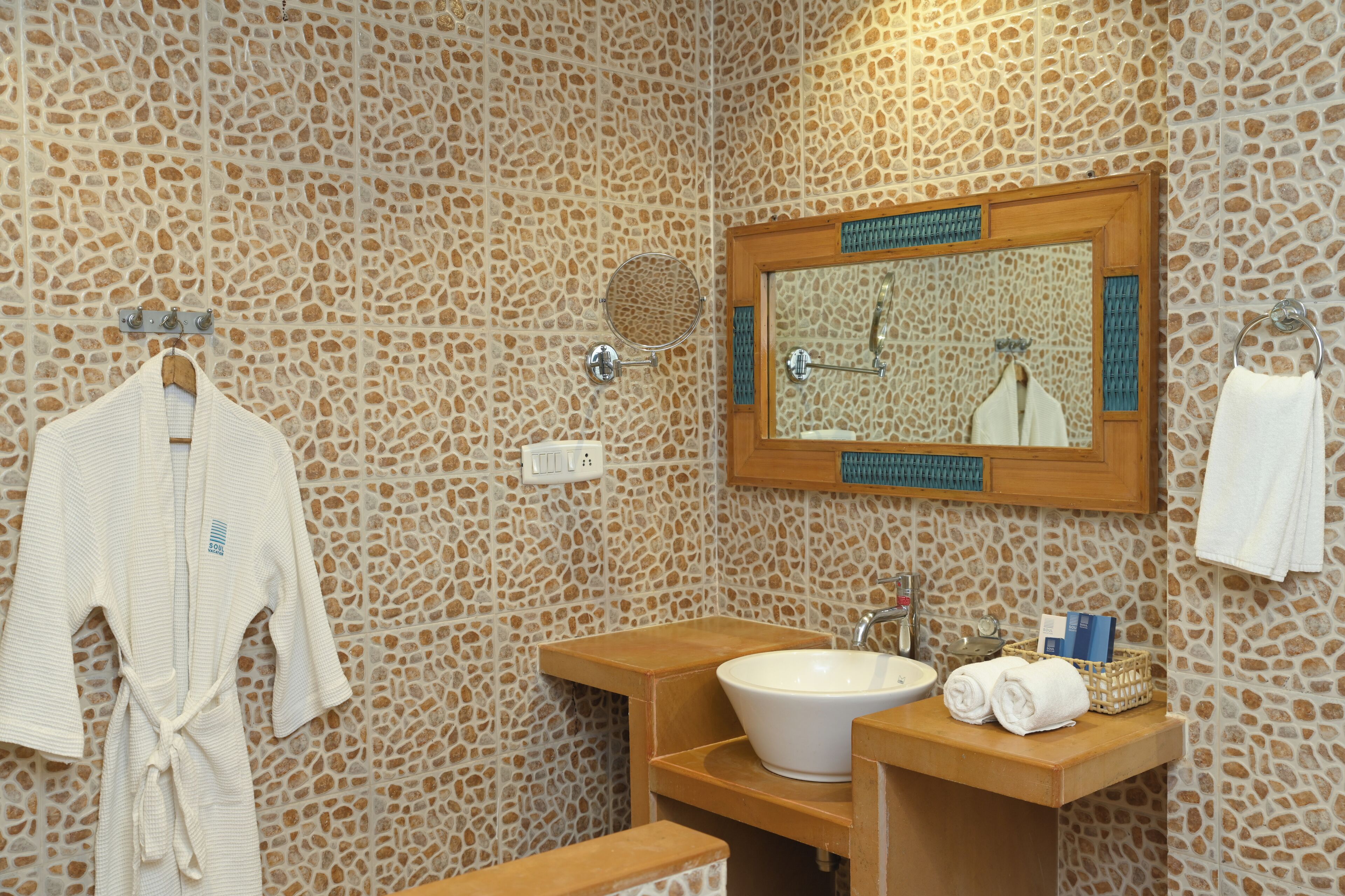 Un baño con pared de diseño de piedra, tocador de madera con lavamanos, espejo y bata colgada en un perchero.