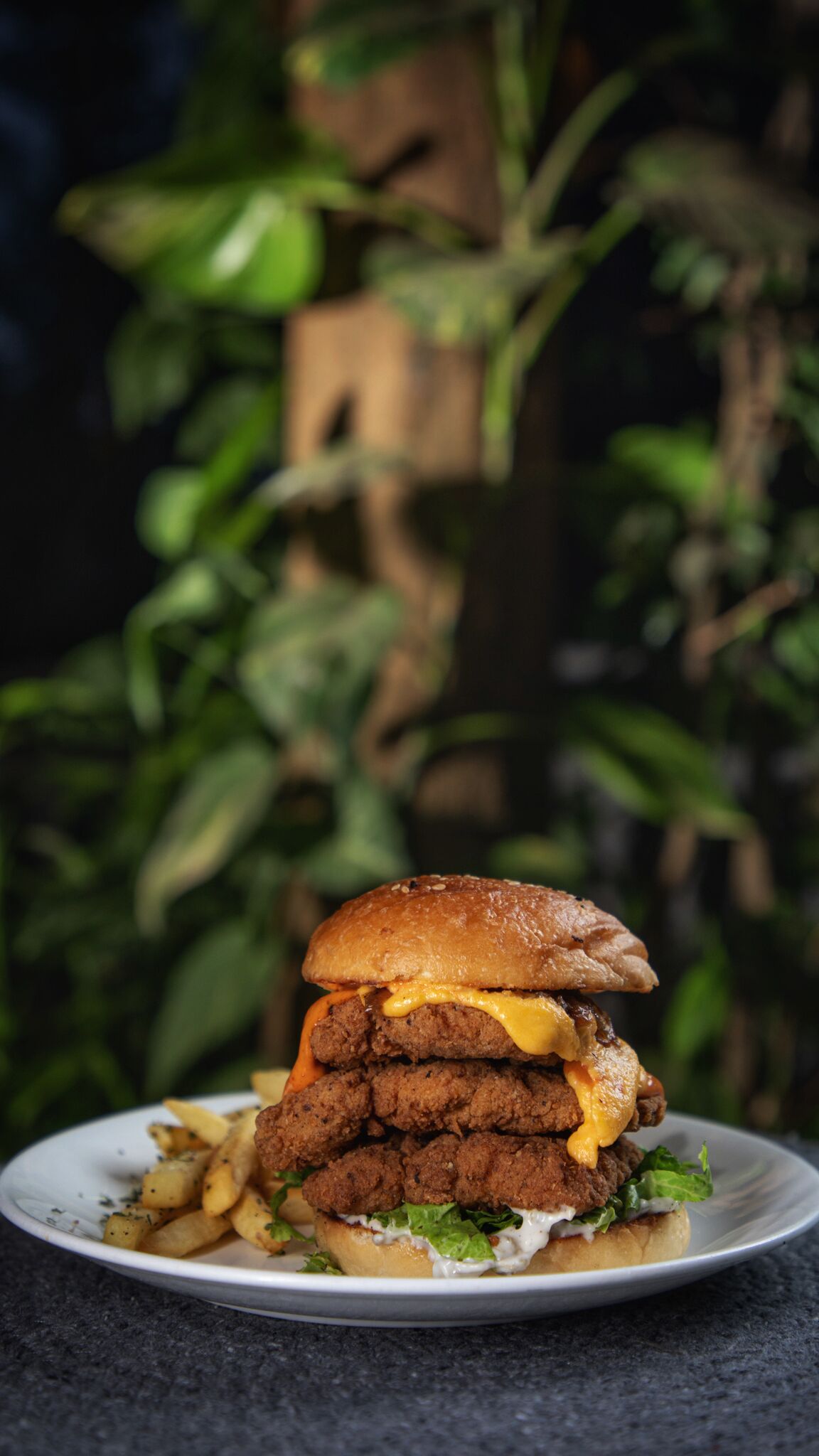 Hamburguesa con varias hamburguesas, queso y lechuga, sobre un plato, con un fondo verde desenfocado.