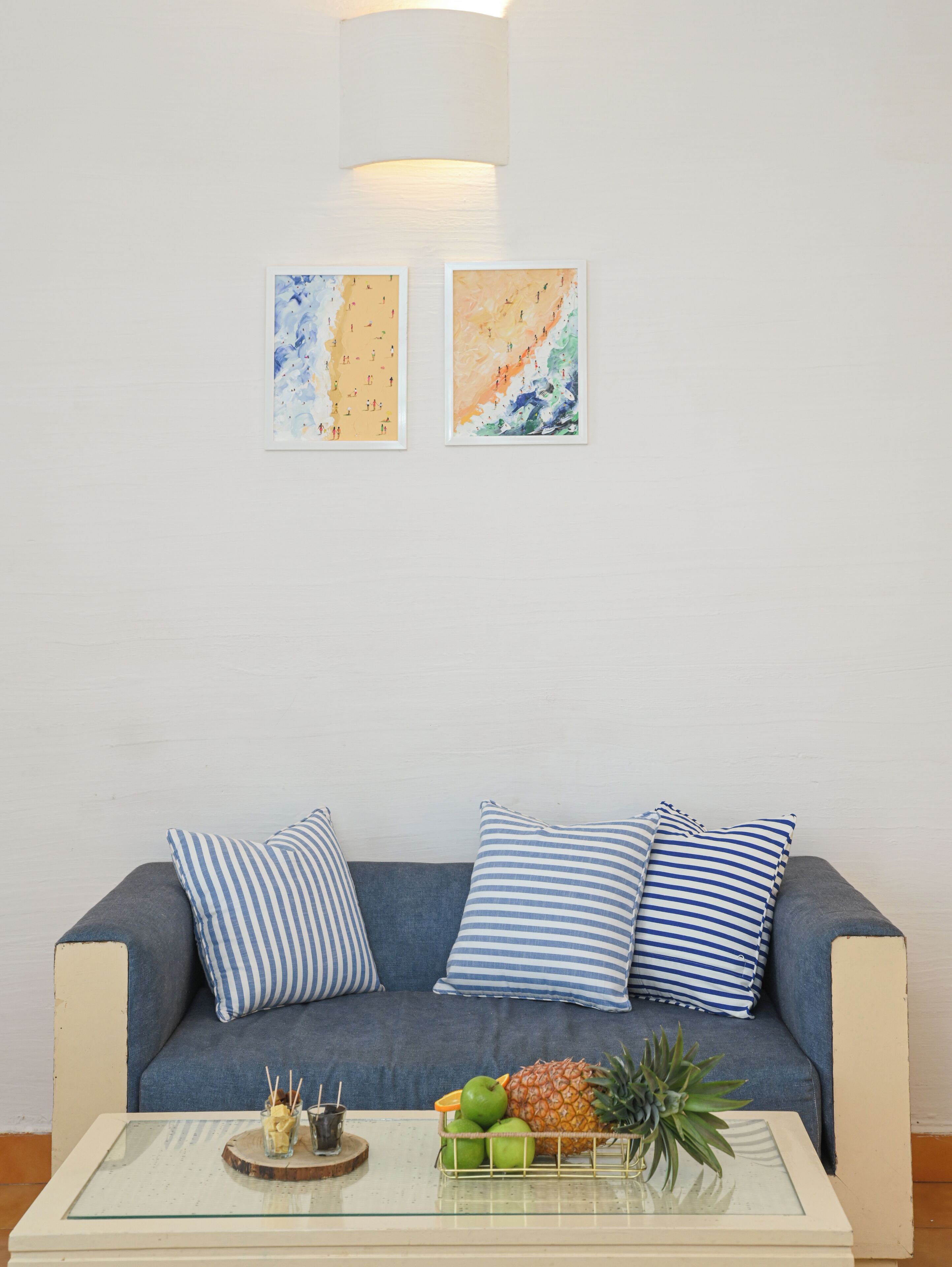 Una sala moderna con un sofá azul, cojines a rayas y una mesa de centro de cristal con fruta y una bebida. Dos obras de arte abstracto decoran la pared.