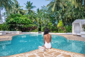 Una mujer sentada junto a una piscina con borde de azulejos, rodeada de exuberante follaje y palmeras.
