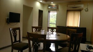 A dining area with a round table, six wooden chairs, a television mounted on the wall, a window with curtains, and an air conditioning unit. - Hotel Sea Lord (Mumbai)