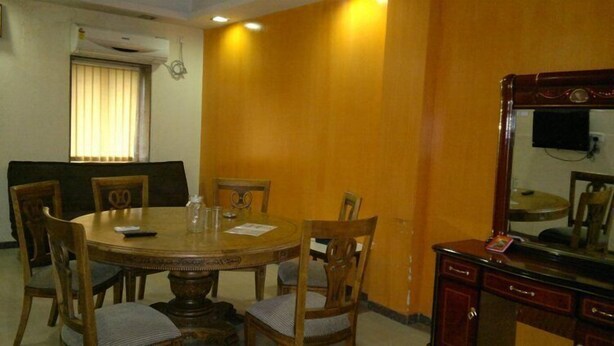 A dining area with a round wooden table, six chairs, a wooden cabinet with a mirror, and a window with blinds. - Hotel Sea Lord (Mumbai)