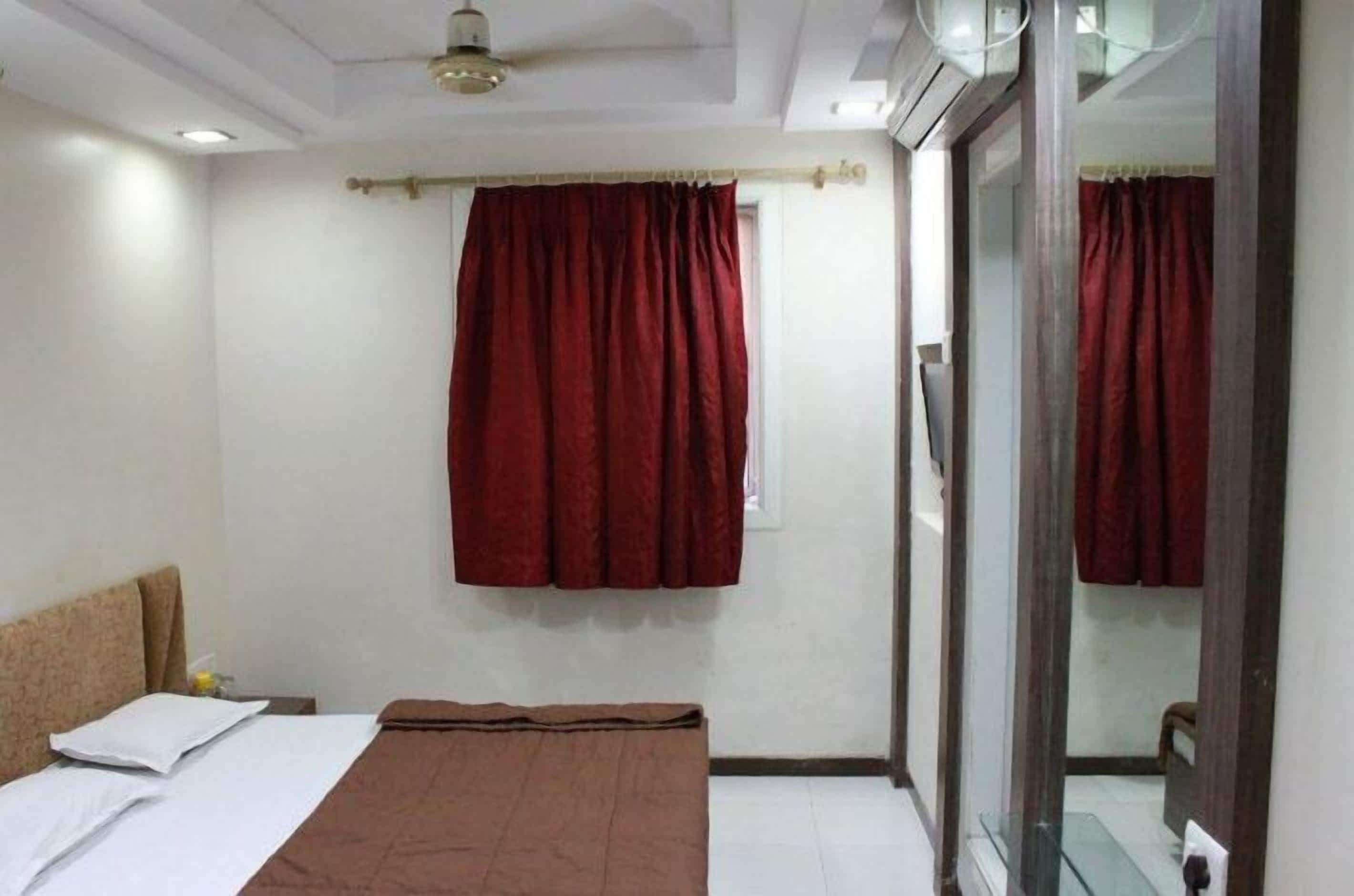 Un dormitorio con cama, ventana con cortinas rojas, ventilador de techo y puerta de vidrio que da a otra habitación.