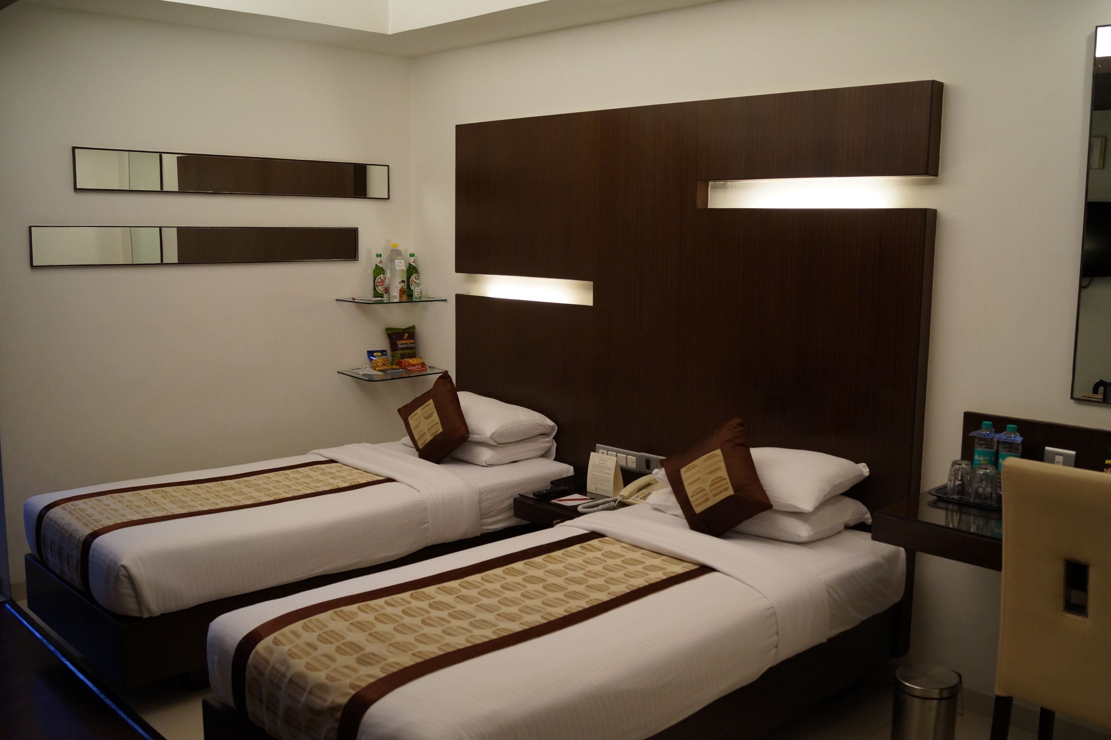 A hotel room with two beds, a wooden headboard, a small shelf with toiletries, and a mirror on the wall.
