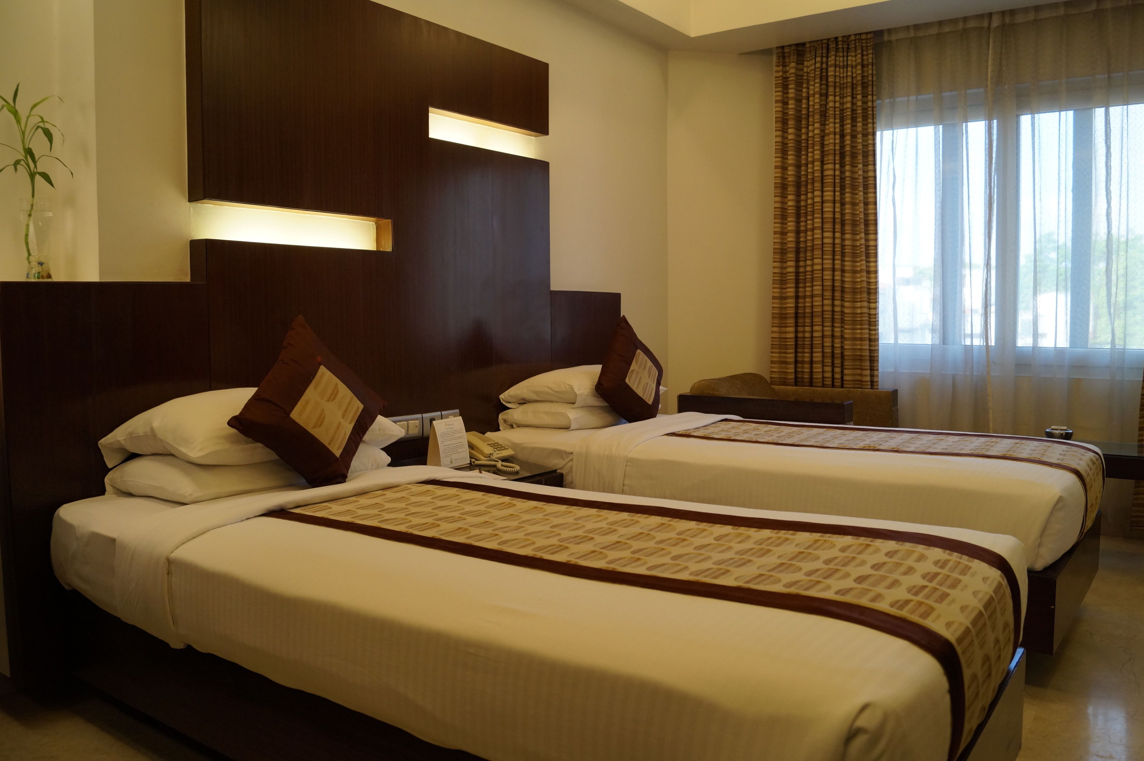 Habitación de hotel con dos camas, una mesita de noche con lámpara, una ventana con cortinas y un aplique de pared.
