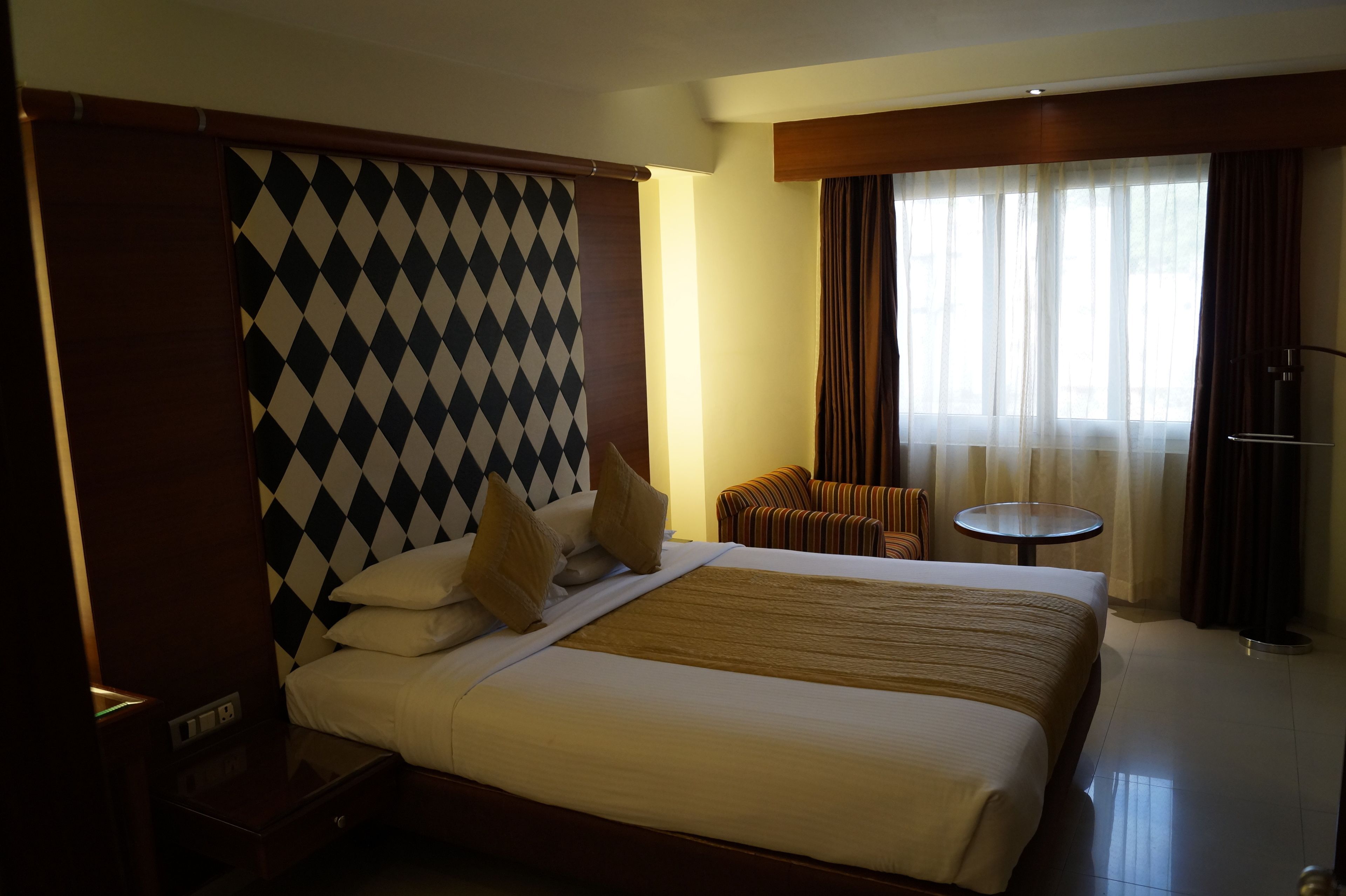 Habitación de hotel con una cama grande, una mesita redonda pequeña, una silla y vistas al exterior a través de cortinas translúcidas.