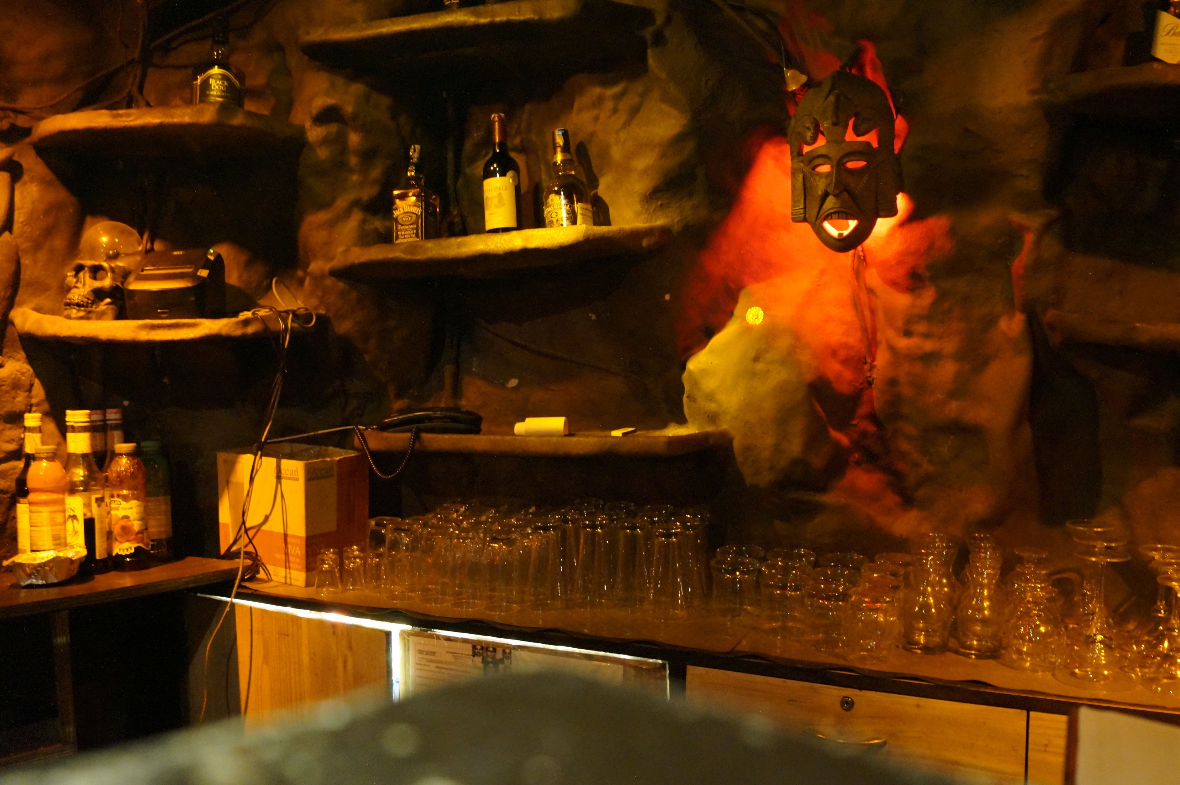 A bar area with shelves of bottles, glasses, and a mask hanging from the wall.