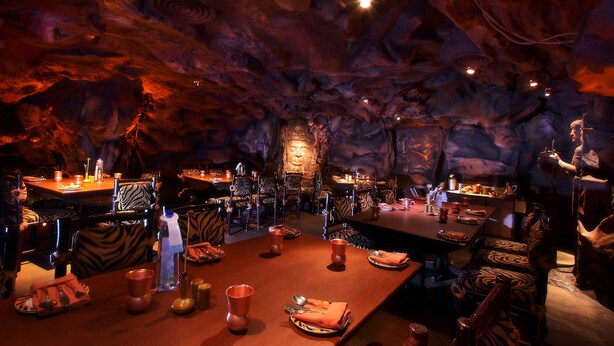 Un ristorante con sedie e tavoli a righe zebrate, apparecchiati per la cena, con interni simili a una grotta, formazioni rocciose e un altare centrale.