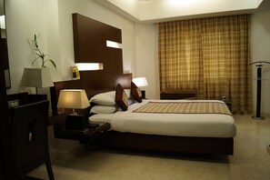 A hotel room with a large bed, two bedside tables with lamps, a nightstand, a wooden headboard, a potted plant, and a window with curtains. - The President Hotel (Bengaluru)