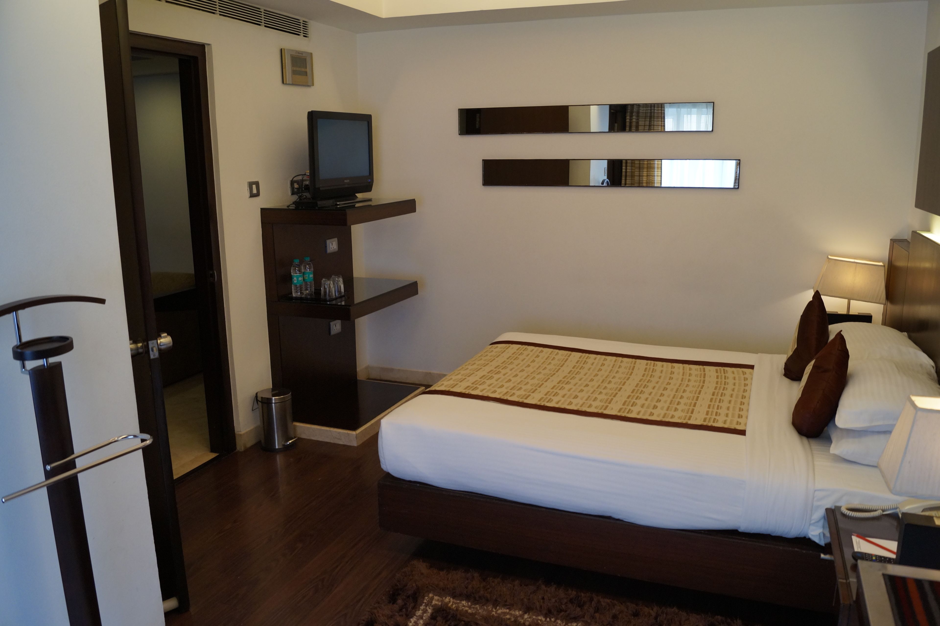 Habitación de hotel con cama, televisor sobre un mueble, un estante con botellas y un cubo de basura.