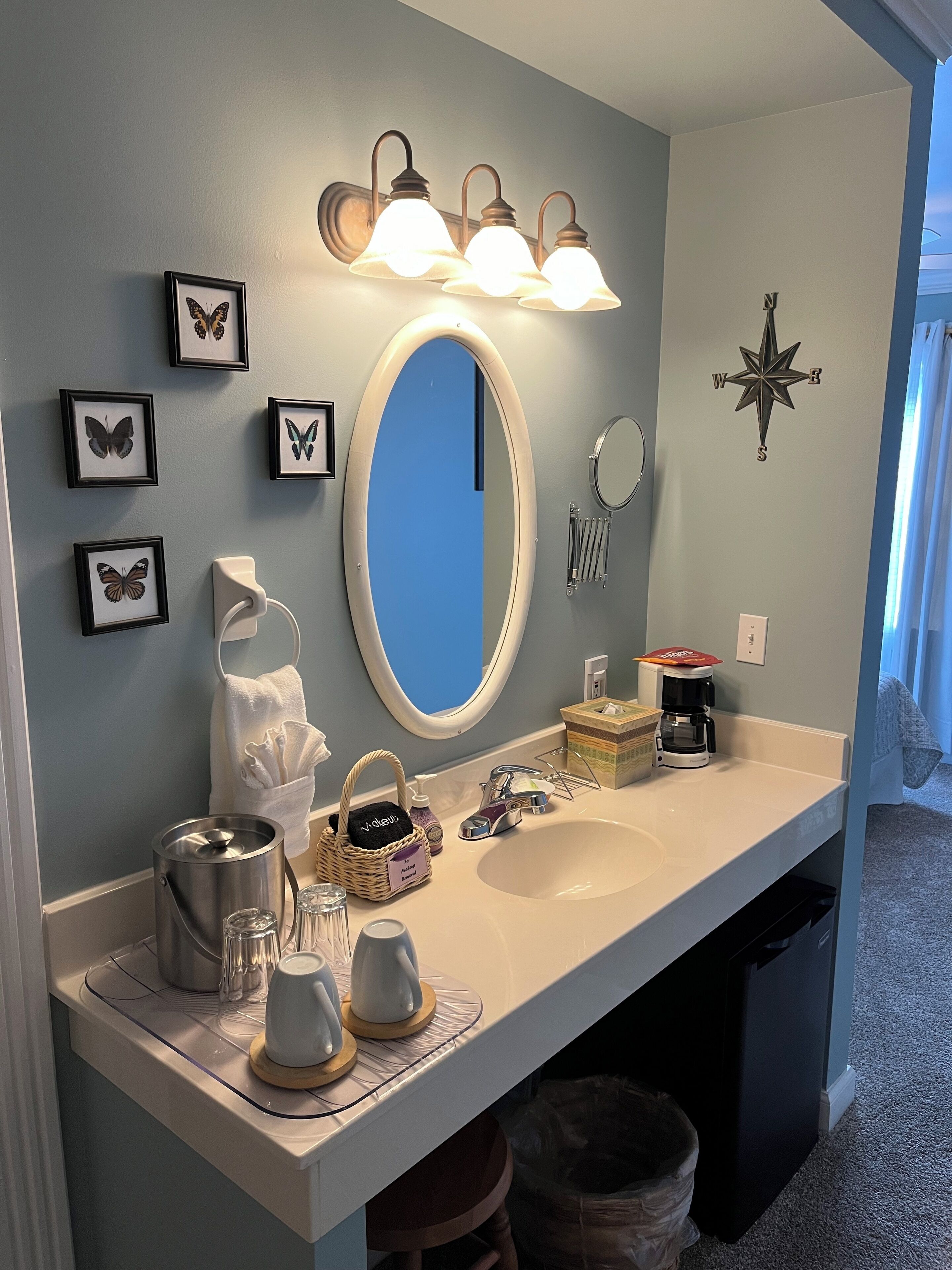 A bathroom with a white vanity, a large mirror, and wall-mounted lights. There are framed butterfly pictures, a compass decoration, and a coffee maker on the counter.