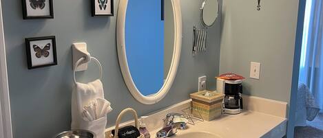 A bathroom with a white vanity, a large mirror, and wall-mounted lights. There are framed butterfly pictures, a compass decoration, and a coffee maker on the counter.