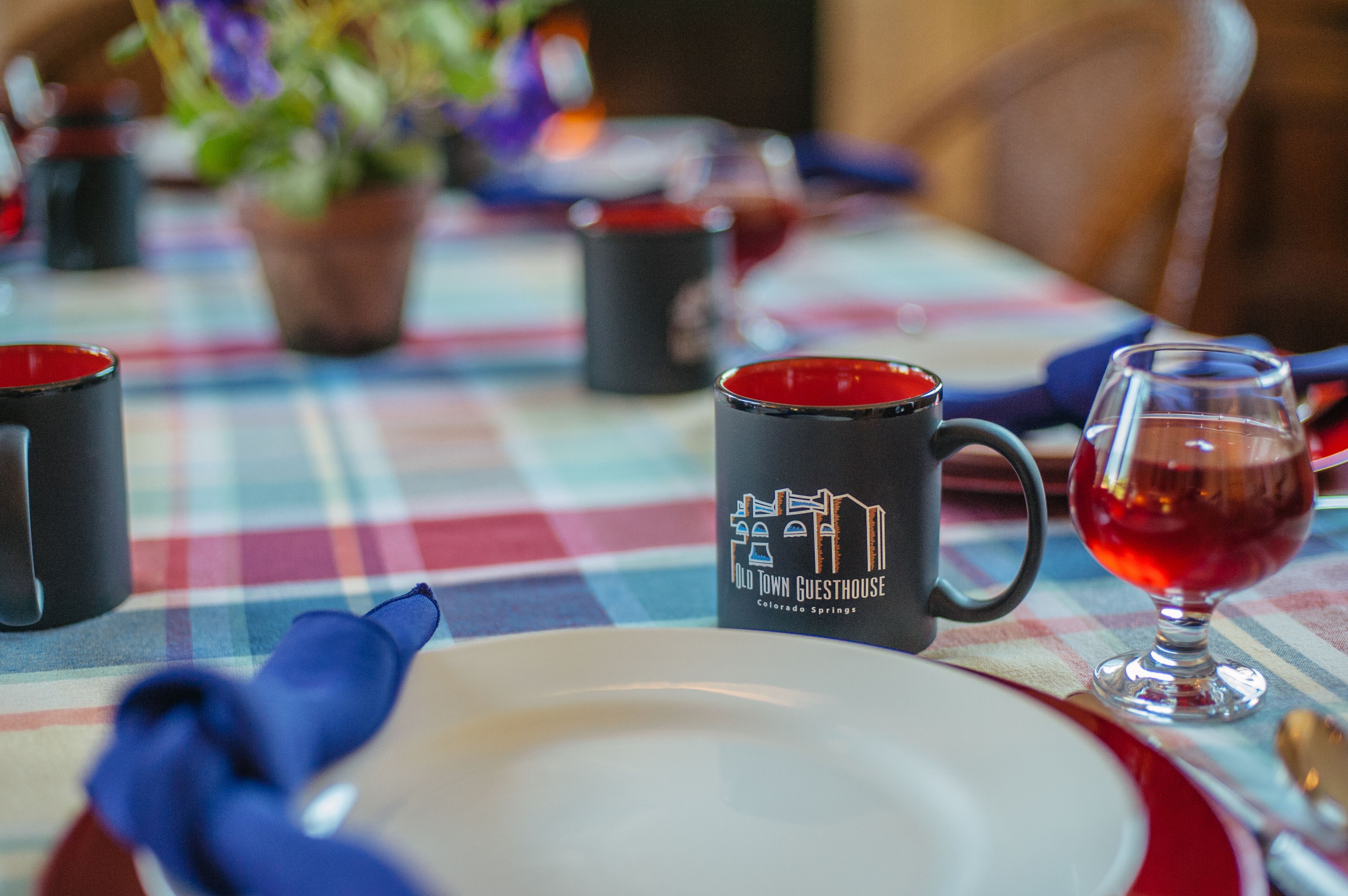 Una mesa puesta con mantel escocés, una copa de vino tinto, una taza negra con un logotipo y un plato blanco.