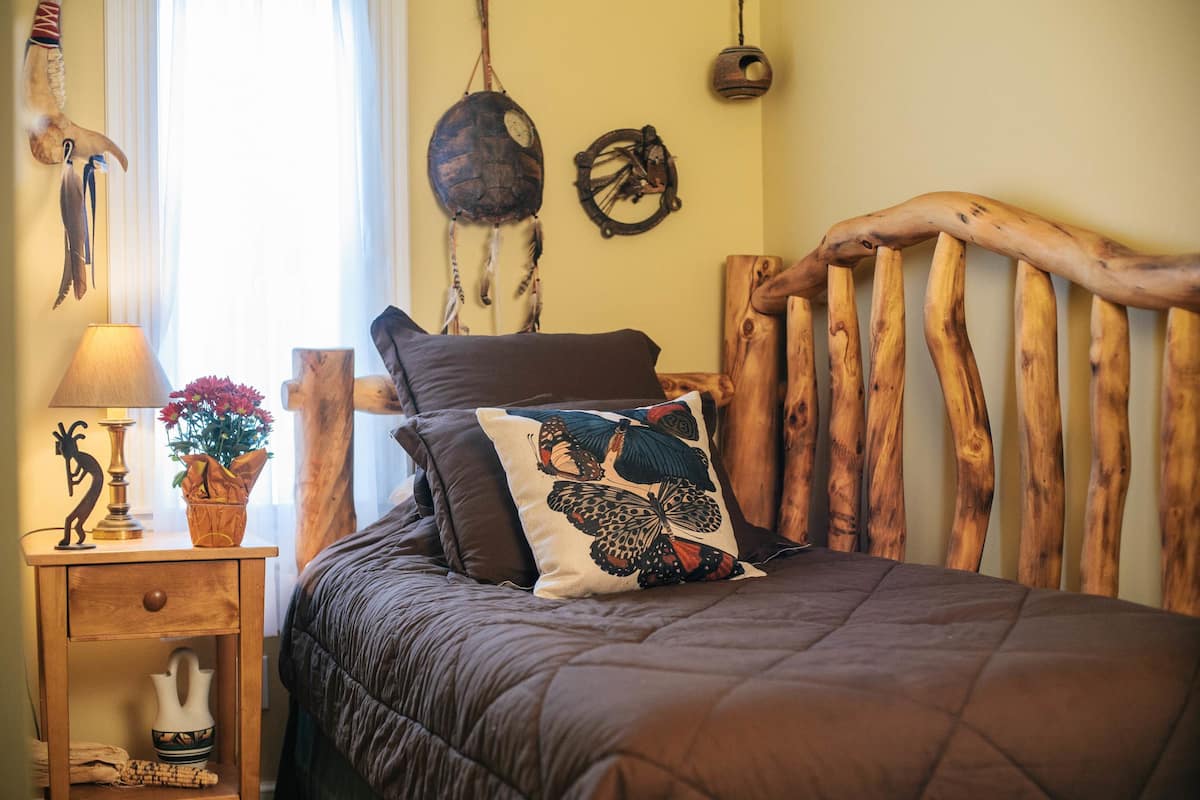 A bed with a wooden headboard, a nightstand with a lamp and decorative items, and wall decorations including a dreamcatcher and a clock.