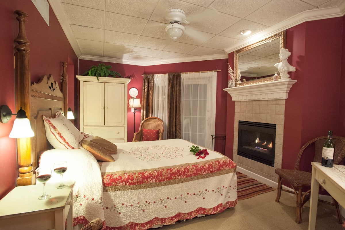 A bedroom with a bed, a fireplace, a mirror, and a nightstand with glasses and a bottle.
