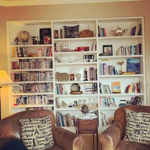A cozy living room with a large bookshelf filled with books, decorative items, and a globe. There are two leather armchairs with travel-themed pillows. - Old Town GuestHouse B&B (Colorado Springs)