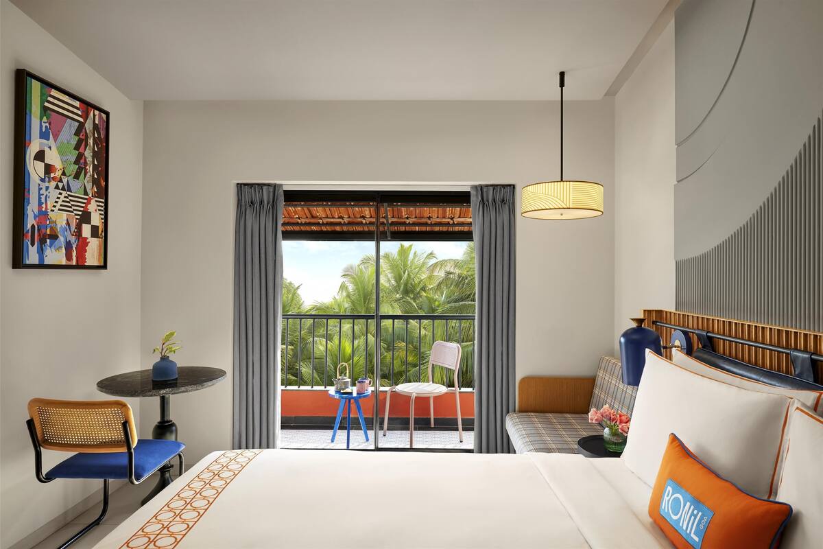 Une chambre d’hôtel avec un grand lit, une petite table avec une chaise, un balcon donnant sur des palmiers et un tableau au mur.