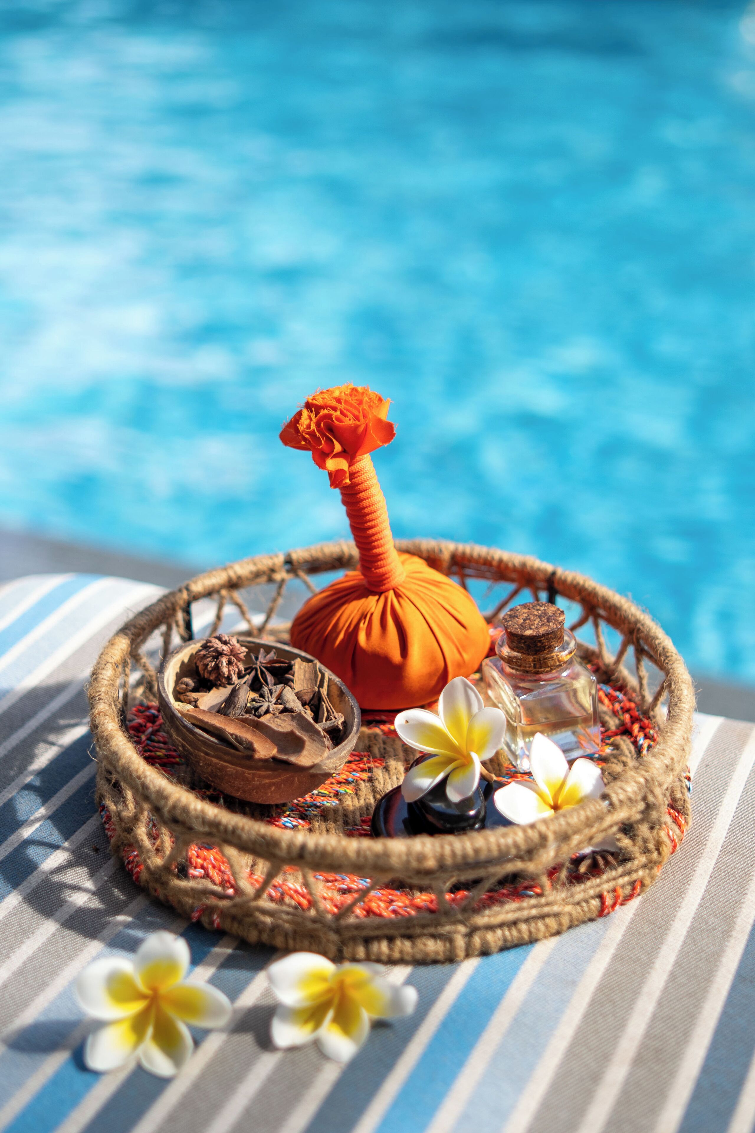 A wicker tray with a massage oil bottle, dried flowers, and a small bottle of essential oil by a poolside.