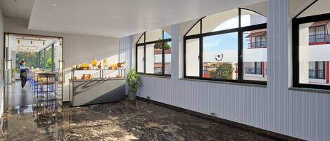 Un moderno vestíbulo de hotel con suelo de mármol, una zona de buffet y amplias ventanas con vistas al exterior.